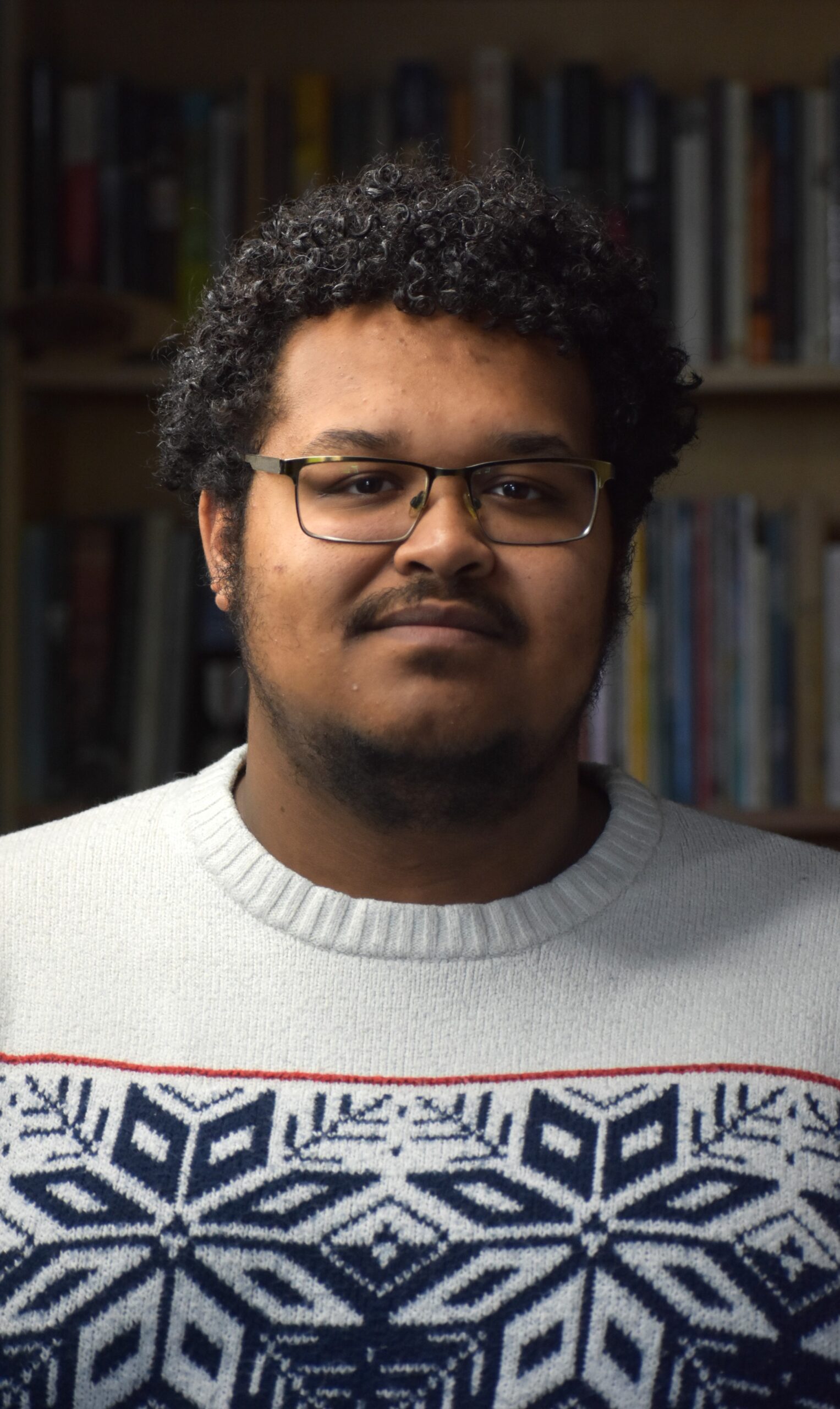 [ID: A portrait of Miguel A. Fenrich with books in the distance behind him. He has curly black hair, black glasses, his light brown skin, a black moustace and light beard, and is wearing a black t-shirt with an open textile-themed dress shirt. He is smiling. End.]