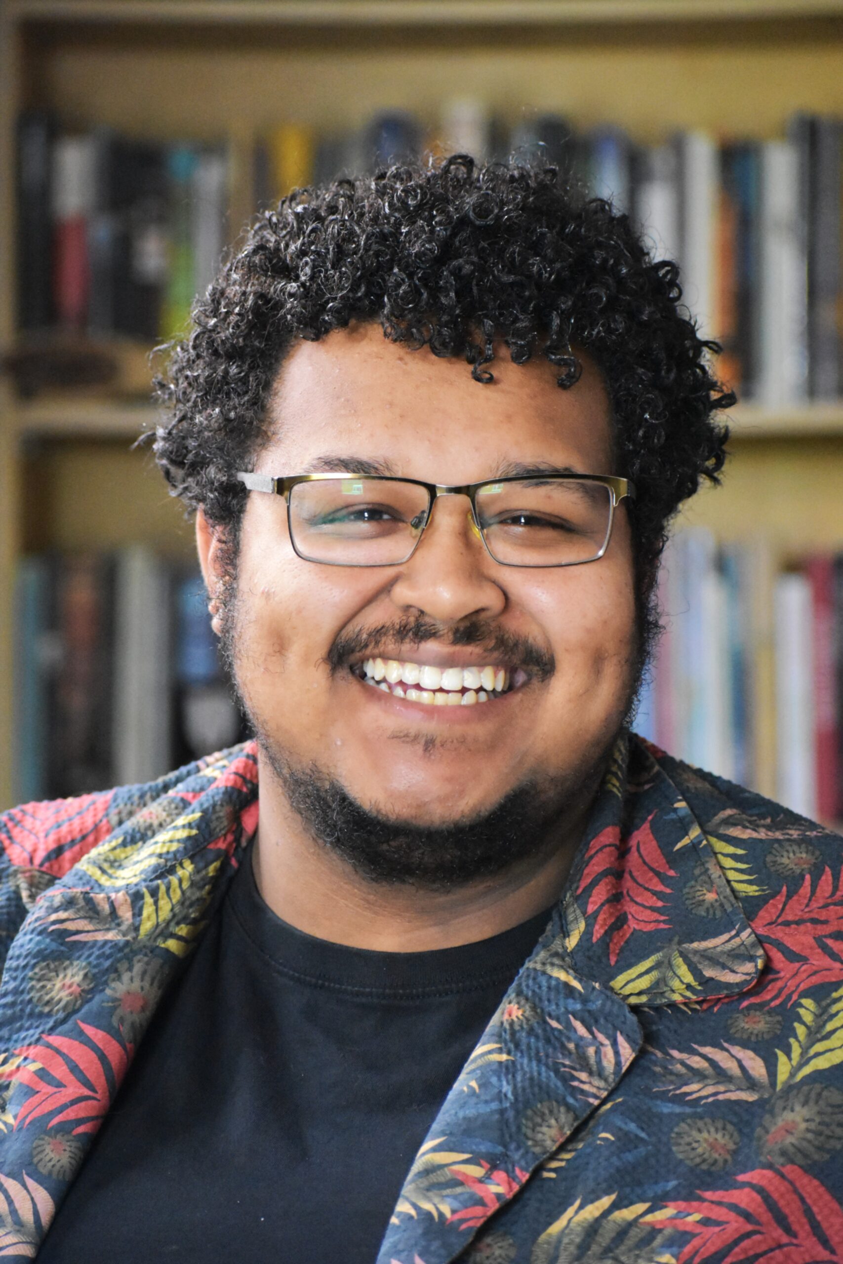 [ID: A portrait of Miguel A. Fenrich with books in the distance behind him. He has curly black hair, black glasses, his light brown skin, a black moustace and light beard, and is wearing a black t-shirt with an open textile-themed dress shirt. He is smiling. End.]